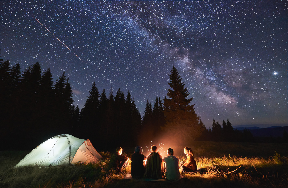 Several young people sitting around a campfire at night with their tent nearby.
